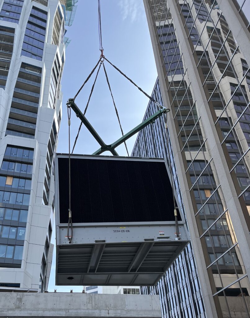A jobsite photo of a large chiller unit being lifted by a crane, including an X shaped lifting beam and wire rope slings.