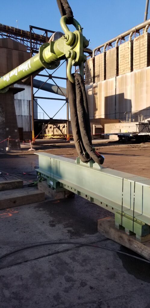 A photo showcasing a modular lifting spreader beam, with a polyester round sling attached to another lifting beam underneath it on a jobsite.