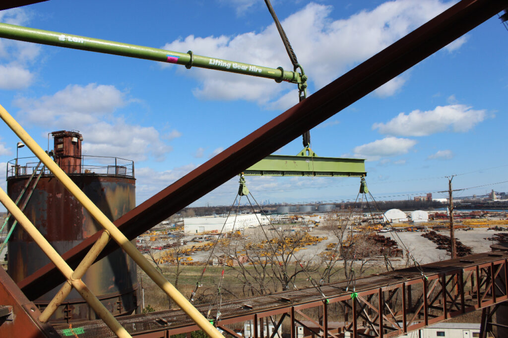 A jobsite photo demonstrating an under-the-crane rigging configuration with modular spreader beam and a lifting beam attached underneath it.