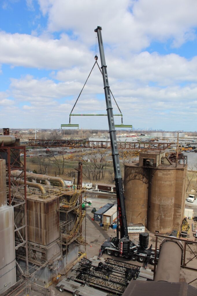 Modular lifting spreader beam with two traditional lifting beams below, attaching by slings, under the hook of a crane with Chicago skyline