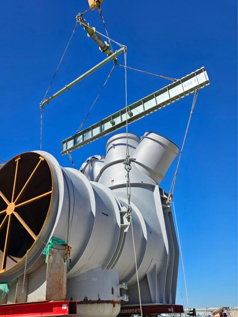 This jobsite photo demonstrates a below-the-hook rigging configuration for a steam duct lift, including wire rope slings, turnbuckles, modular spreader beams, and a 100-ton multi-lug lifting beam.