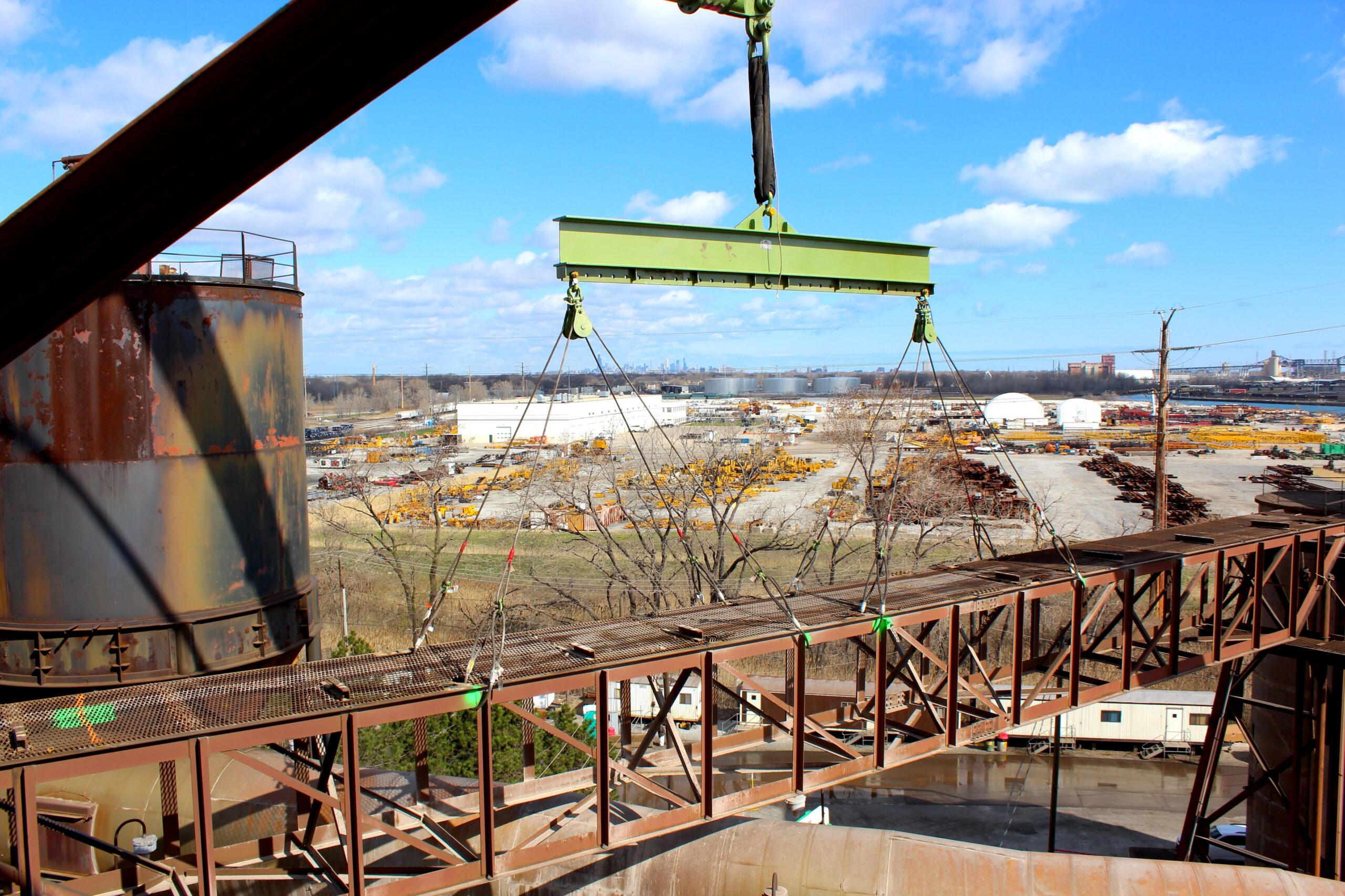 A jobsite photo of a crane lifting a bridge section with below-the-hook rigging from LGH, including a lifting beam, round slings, sheave blocks, wire rope slings, and shackles.